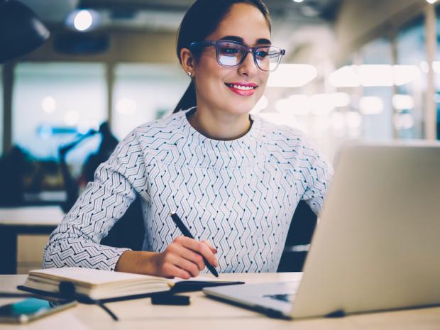 a smiling woman holding a pen looking at her laptop screen